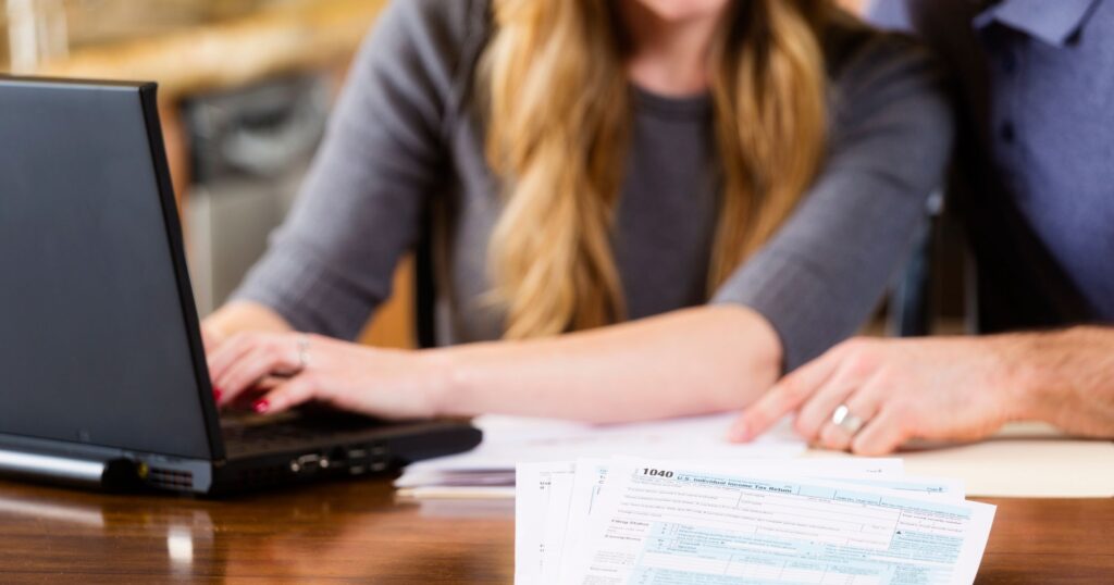 Two residents preparing taxes together at a kitchen table using a laptop, with 1040 tax forms in the foreground.