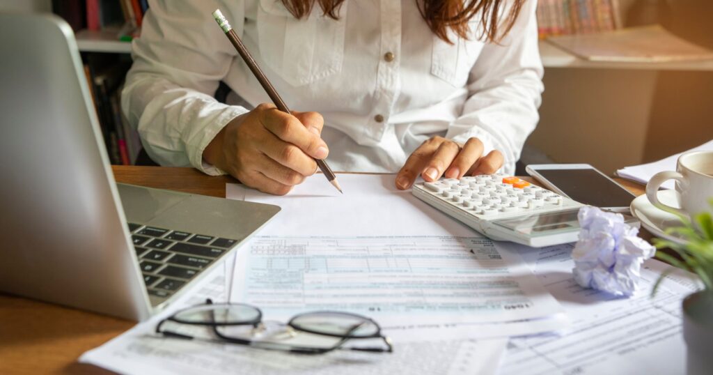 Person in white shirt working on financial documents at a desk with a laptop, calculator, pencil, and cup of coffee.