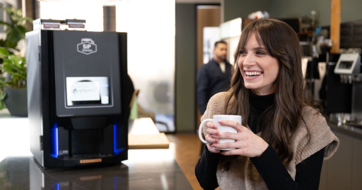 female stands near sipwithme coffee amenity machine drinking a cup of coffee smiling