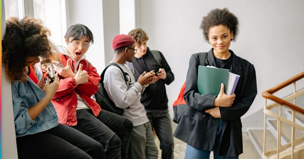 Student holding notebooks walking through a student housing building while other students use phones in the background, highlighting different student needs and daily routines