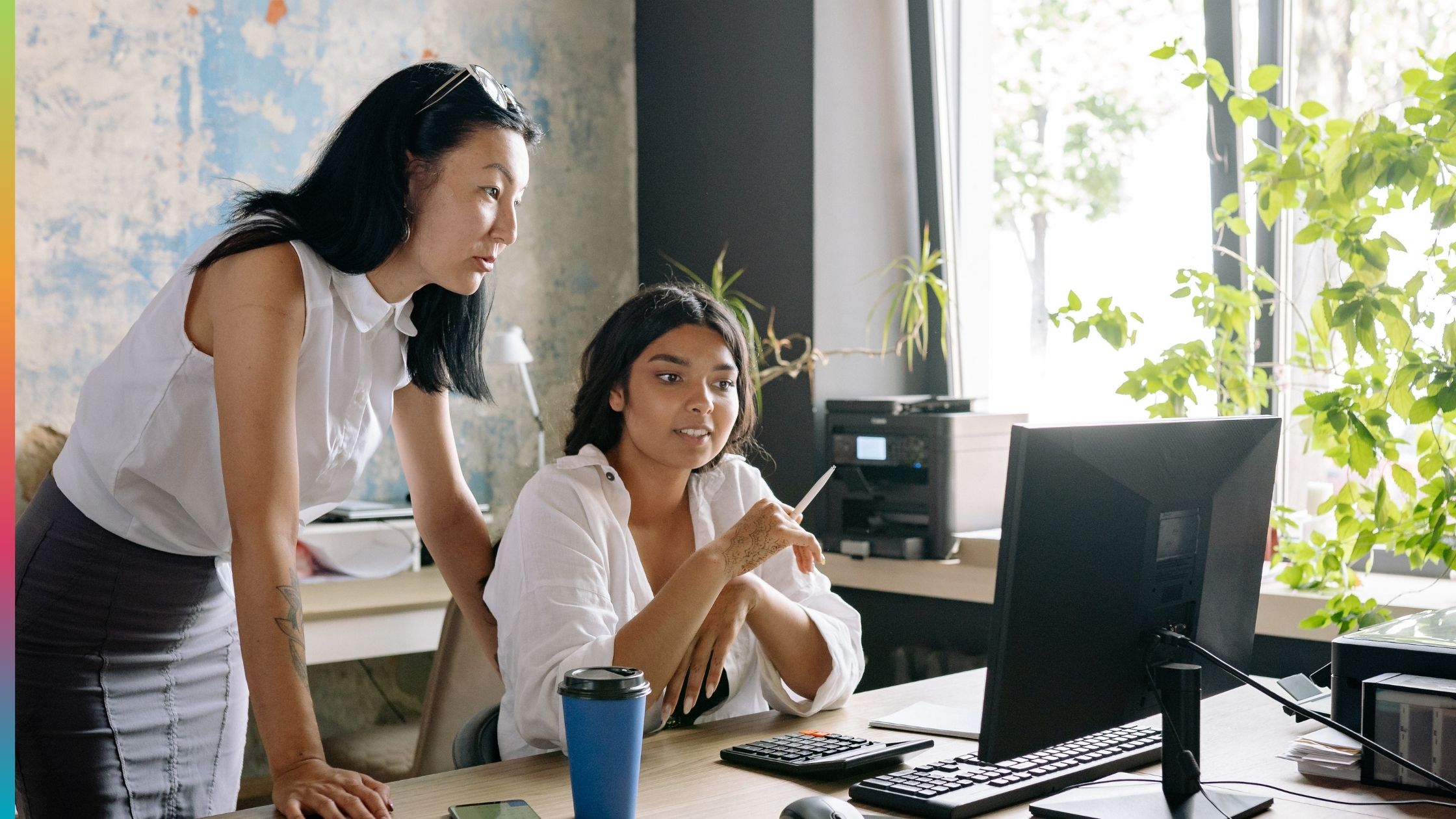 Two office employees reviewing information on a computer with an office printer in the background.